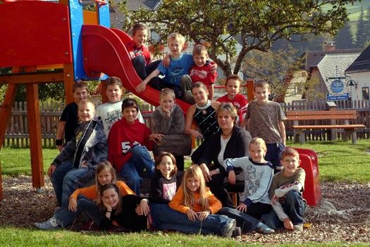 Eine Gruppe von Kindern und zwei Erwachsenen posiert für ein Foto auf einem Spielplatz mit einer Rutsche und Bäumen im Hintergrund.