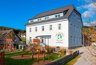 Ein großes weißes Gebäude mit einem Spielplatz davor. Das Gebäude hat ein graues Dach und mehrere Fenster. Ein Logo befindet sich an der Wand.