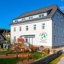 Ein weißes Gebäude mit grauem Dach steht auf dem Land. Es hat mehrere Fenster und ein Logo an der Wand. Vor dem Gebäude befindet sich ein Spielplatz mit einer Rutsche und Klettergerüst.
