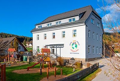 Ein weißes Gebäude mit grauem Dach steht auf dem Land. Es hat mehrere Fenster und ein Logo an der Wand. Vor dem Gebäude befindet sich ein Spielplatz mit einer Rutsche und Klettergerüst.