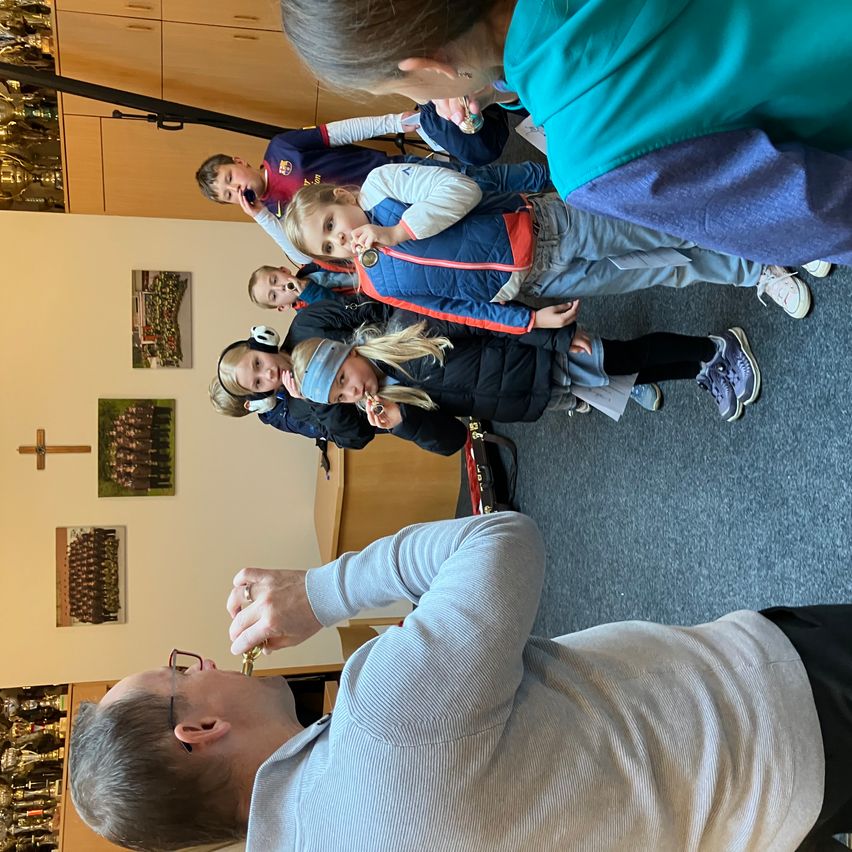 Eine Gruppe von Kindern mit Kopfhörern steht im Kreis mit einem erwachsenen Mann, der ein goldenes Objekt hält. Sie befinden sich in einem Raum mit einem Holzschrank, einem Kreuz an der Wand und Bildern.