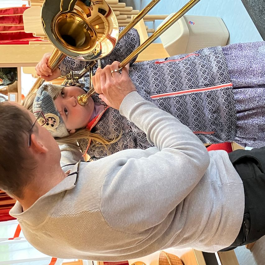 Ein Mann mit einer Brille hilft einem jungen Mädchen in einem grauen Mantel und Hut, ein Posaune zu spielen.