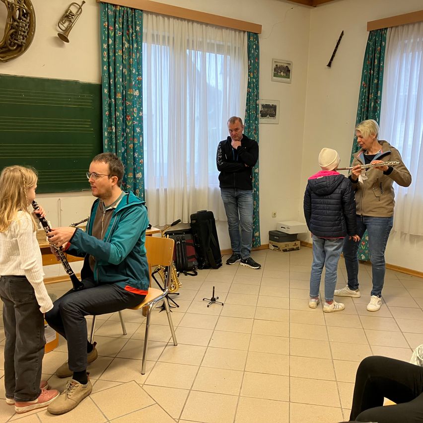 In einem Klassenzimmer unterrichtet ein Mann ein Mädchen im Klarinettenspiel. Eine andere Frau lehrt ein Kind das Flötenspiel. Zwei Erwachsene stehen in der Nähe.