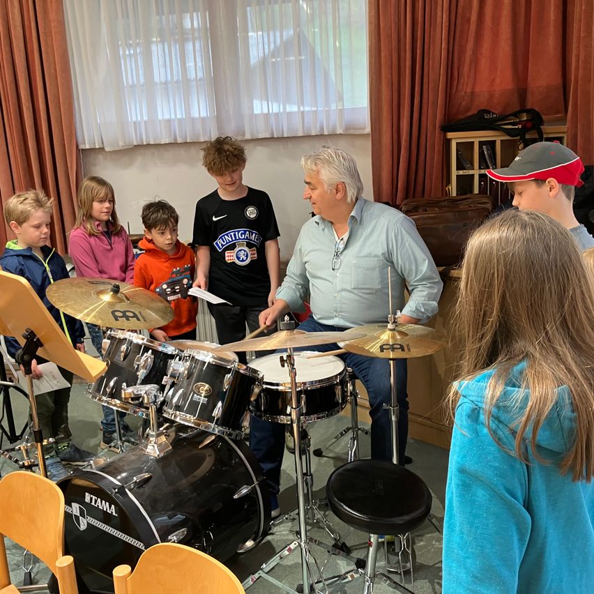 In einem Klassenzimmer spielt ein erwachsener Mann die Drums, während eine Gruppe von Kindern zuschaut. Die Kinder stehen vor den Drums. Der Mann trägt ein blaues Hemd. Die Kinder tragen Jacken und Hosen. Die Drums stehen auf einem Hocker.