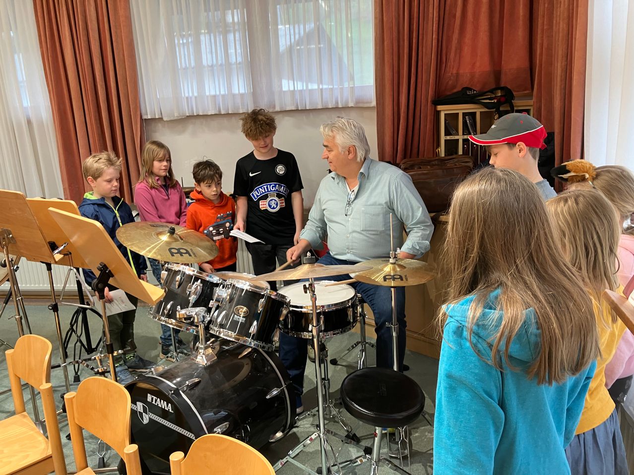In einem Klassenzimmer spielt ein erwachsener Mann die Drums, während eine Gruppe von Kindern zuschaut. Die Kinder stehen vor den Drums. Der Mann trägt ein blaues Hemd. Die Kinder tragen Jacken und Hosen. Die Drums stehen auf einem Hocker.