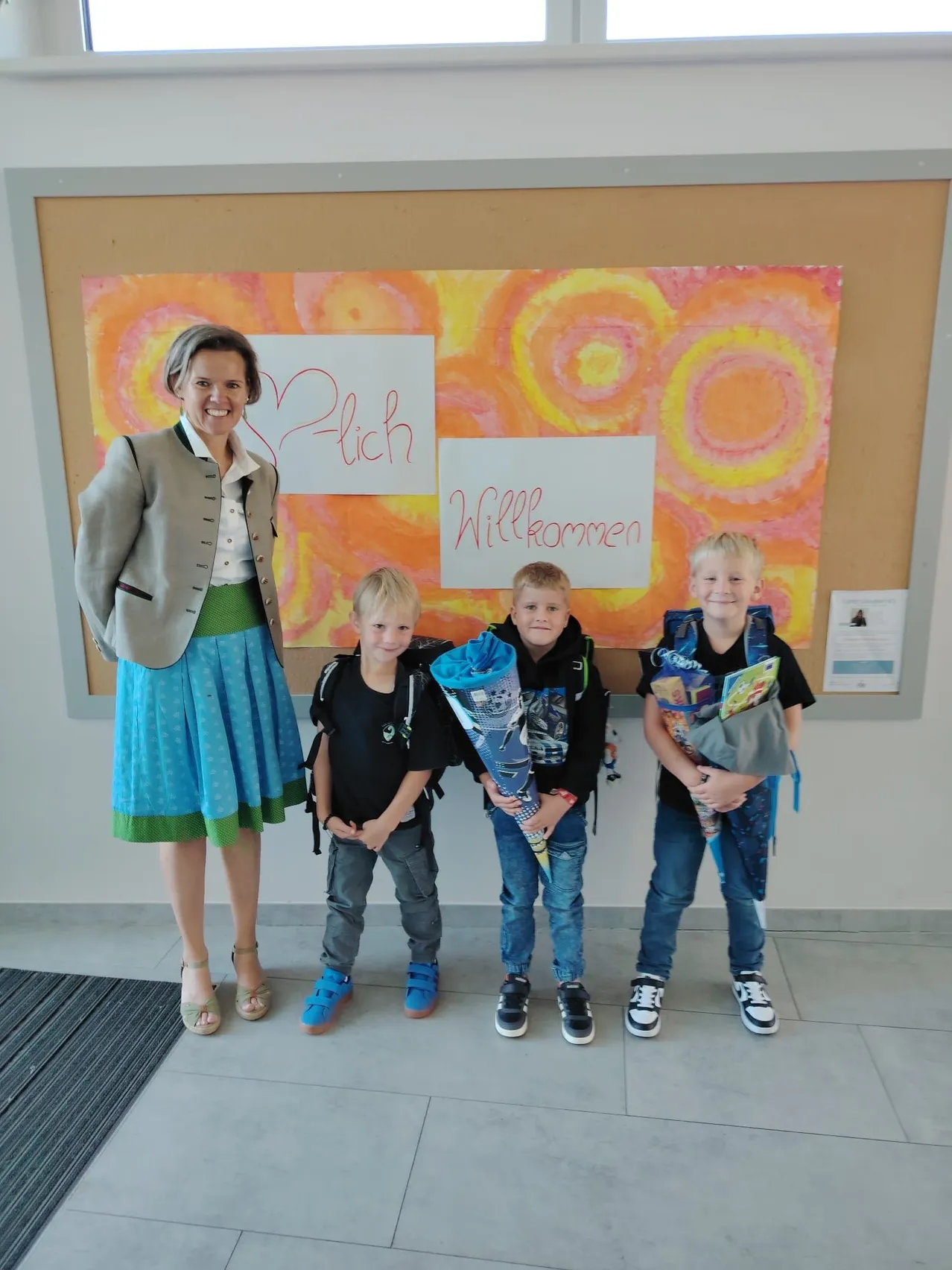 Eine Frau und drei Jungen stehen vor einem Bulletin Board mit einem farbenfrohen Gemälde. Die Frau und die Jungen lächeln. Das Gemälde enthält ein Herz und die Worte 'Plich, Willkommen'.