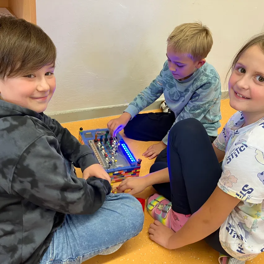 Drei Kinder sitzen auf dem Boden und beschäftigen sich mit einem Lego-Gerät mit blauen Lichtern. Sie lächeln und scheinen ihre Aktivität zu genießen.