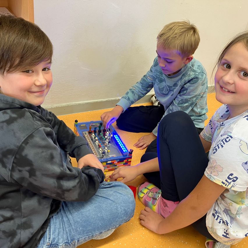 Drei Kinder sitzen auf dem Boden und beschäftigen sich mit einem Lego-Gerät mit blauen Lichtern. Sie lächeln und scheinen ihre Aktivität zu genießen.