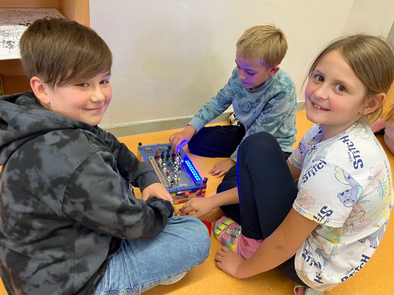 Drei Kinder sitzen auf dem Boden und beschäftigen sich mit einem Lego-Gerät mit blauen Lichtern. Sie lächeln und scheinen ihre Aktivität zu genießen.