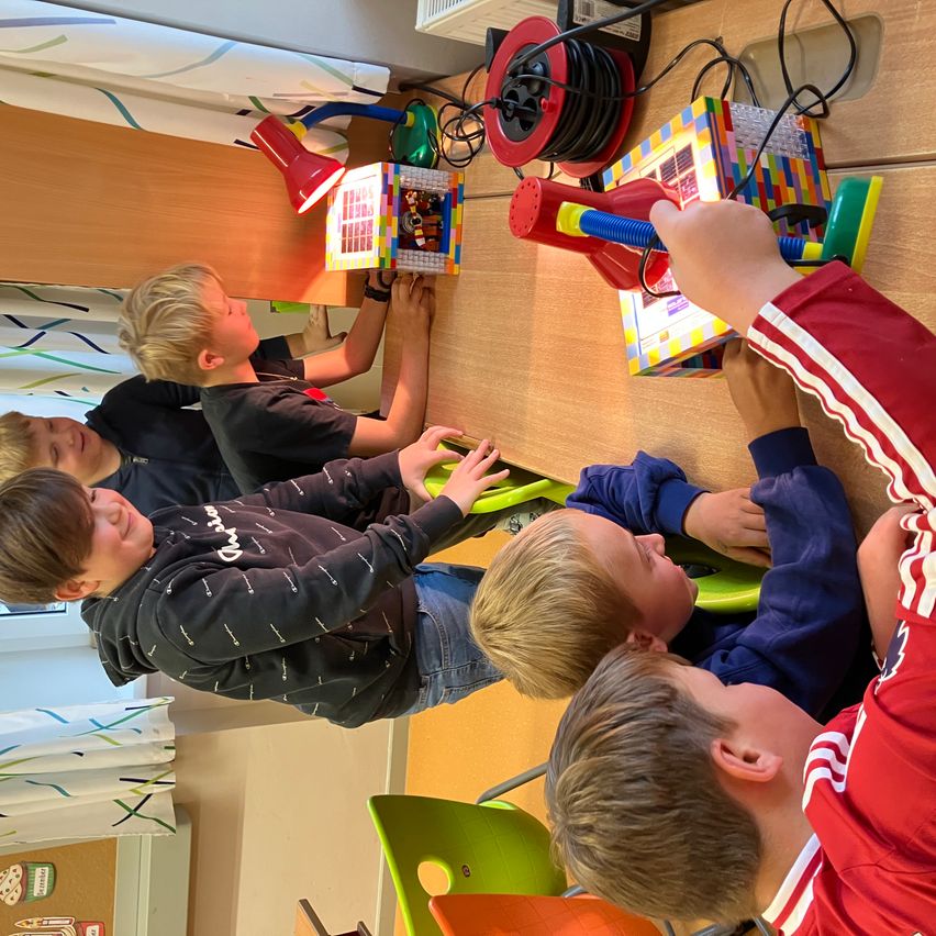 Fünf junge Jungen sitzen um einen Tisch herum und spielen mit LEGO. Jeder Junge konzentriert sich auf verschiedene LEGO-Strukturen, einer mit einer roten Lampe, andere mit bunten Blöcken.