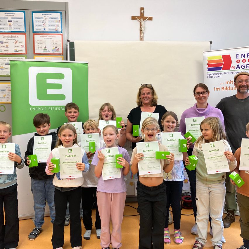 Eine Gruppe von Kindern mit Urkunden steht vor einem grünen Banner. Dahinter lächeln zwei Frauen und ein Mann. Ein Kreuz ist an der Wand befestigt.
