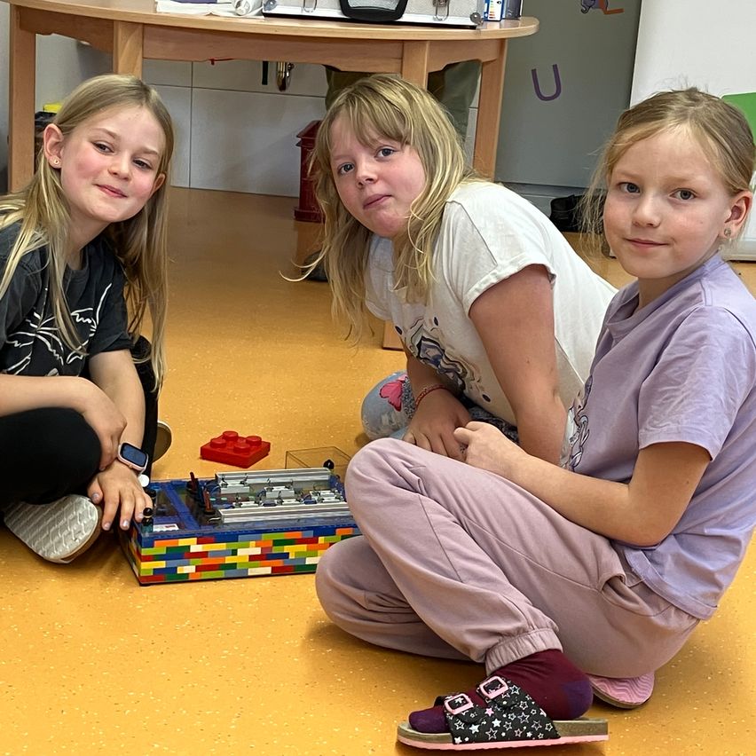 Drei Mädchen sitzen auf dem Boden. Sie sehen sich einen Lego-Bausatz an und scheinen daran zu arbeiten. Sie befinden sich wahrscheinlich in einem Klassenzimmer.