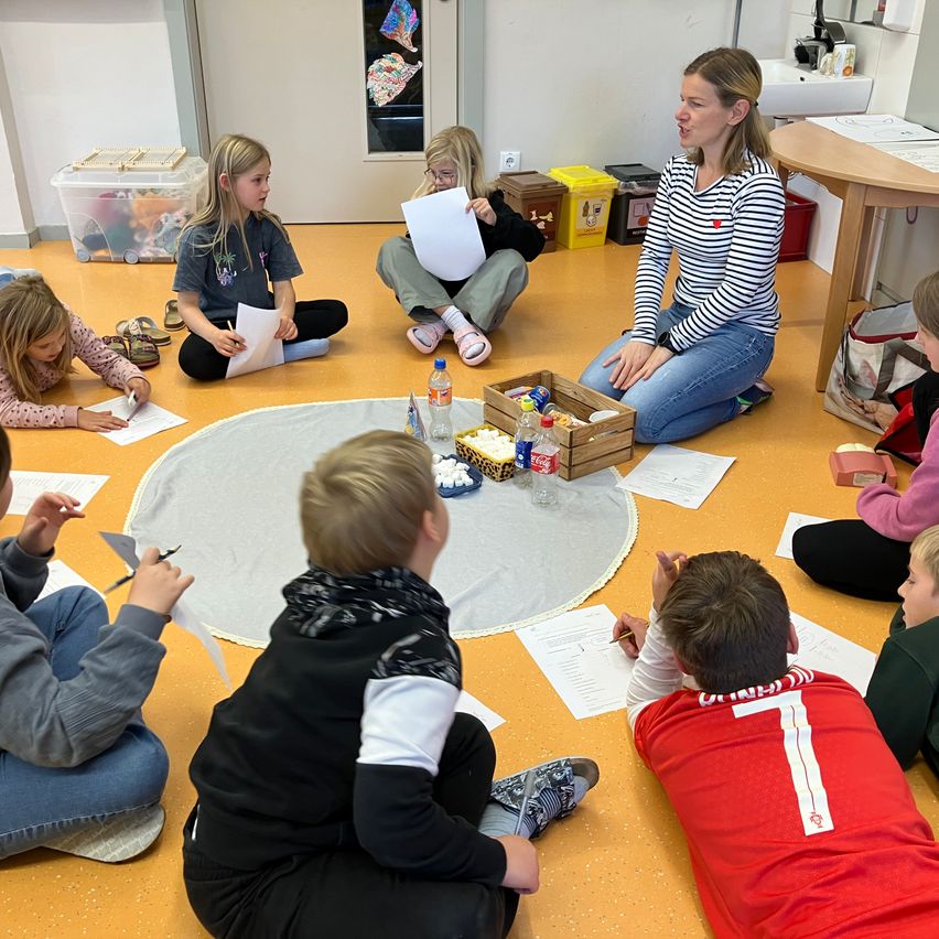 Eine Gruppe von Kindern sitzt im Kreis auf dem Boden mit einer Lehrerin, hält Papiere in den Händen und schaut eine Frau an, die aus einem Papier liest. Der Raum hat gelben Boden, und es gibt verschiedene Gegenstände und einen Tisch im Hintergrund.