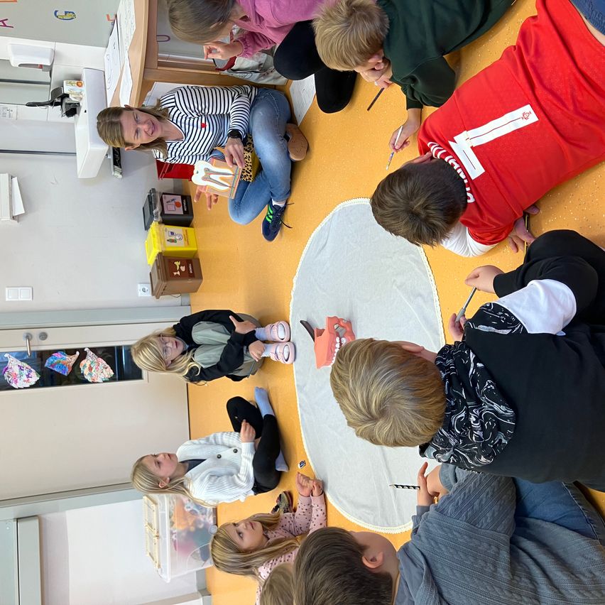 Ein Klassenzimmer mit Kindern, die in einem Kreis um eine weiße Matte herum sitzen, wobei ein Kind ein Zahnmodell hält. Die Lehrerin sitzt in der Mitte, lächelt und hält ein Zahnmodell.