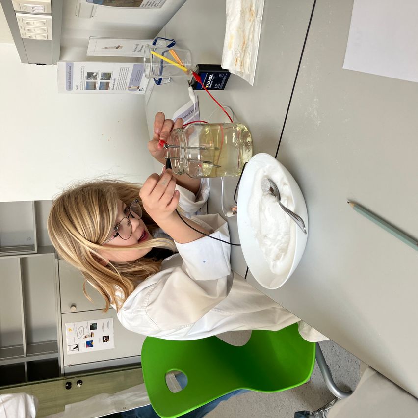 Ein junges Mädchen in einer weißen Laborkittel führt ein wissenschaftliches Experiment durch, hält ein mit Drähten verbundenes Glas und eine Platte mit einer Substanz. Sie sitzt an einem Tisch mit verschiedenen Gegenständen darauf.