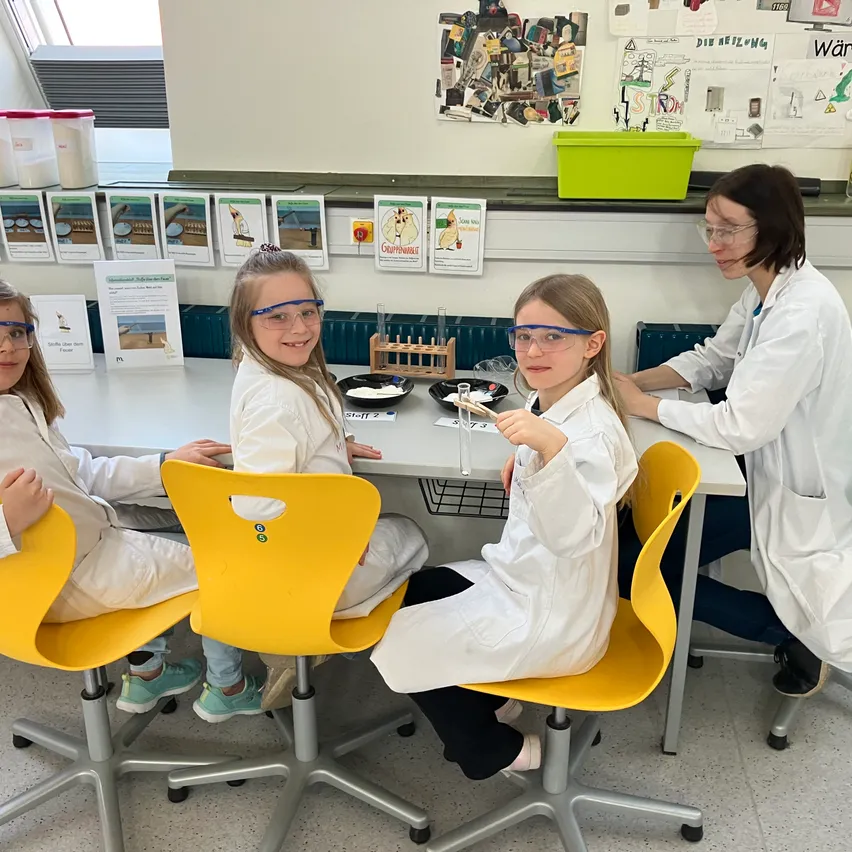 Drei Mädchen in weißen Laborkitteln sitzen um einen Tisch in einem Klassenzimmer herum und tragen Schutzbrillen. Eine Frau in einem Laborkittel steht hinter ihnen und beaufsichtigt sie. Verschiedene naturwissenschaftliche Materialien sind auf dem Tisch und im Regal hinter ihnen.