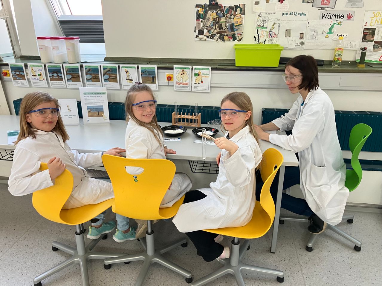 Drei Mädchen in weißen Laborkitteln sitzen um einen Tisch in einem Klassenzimmer herum und tragen Schutzbrillen. Eine Frau in einem Laborkittel steht hinter ihnen und beaufsichtigt sie. Verschiedene naturwissenschaftliche Materialien sind auf dem Tisch und im Regal hinter ihnen.