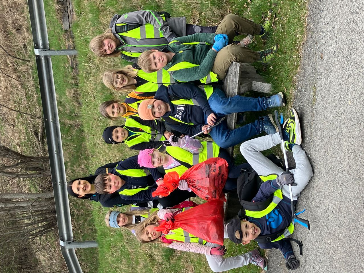 Eine Gruppe von Menschen in Warnwesten sitzt und steht auf dem Rasen. Sie haben Müllsäcke und Stöcke. Sie lächeln und posieren für ein Foto.