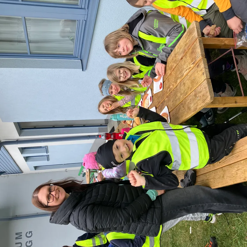 Eine Gruppe von Kindern in Warnwesten sitzt draußen auf einer Bank, einige lächeln und essen. Eine Frau steht neben ihnen, ebenfalls lächelnd. Hinter ihnen ist ein Gebäude mit Fenstern und einem Schild mit der Aufschrift 'UM GG' zu sehen.