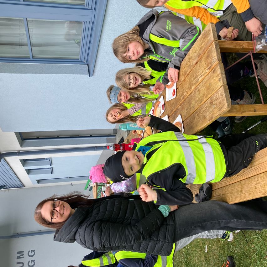 Eine Gruppe von Kindern in Warnwesten sitzt draußen auf einer Bank, einige lächeln und essen. Eine Frau steht neben ihnen, ebenfalls lächelnd. Hinter ihnen ist ein Gebäude mit Fenstern und einem Schild mit der Aufschrift 'UM GG' zu sehen.