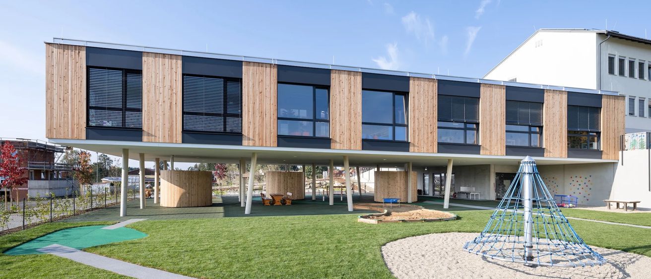 Ein modernes Gebäude mit Holzfassaden, großen Fenstern und einem darunterliegenden Spielplatz. Der Spielplatz beinhaltet eine Rutsche und einen Sandkasten.