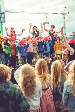 Eine Gruppe von Kindern und Erwachsenen steht mit Bauhelmen auf der Bühne. Einige von ihnen heben die Hände. Die Kinder tragen verschiedene Jacken und Hosen.