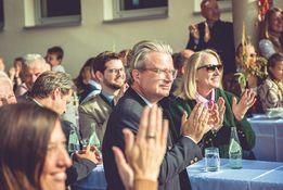 Eine Versammlung von Menschen in einem Raum, die applaudieren. Ein Mann mit Brille lächelt. Eine Frau in einer grünen Jacke klatscht. Es gibt Tische mit Flaschen und Gläsern.