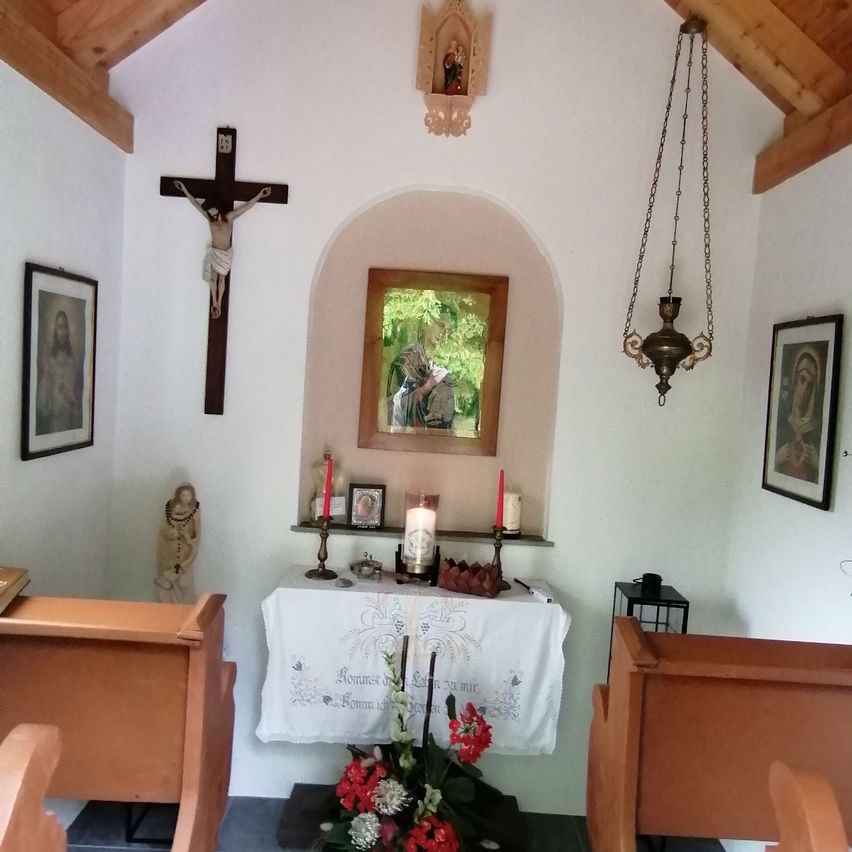 Eine kleine Kapelle mit einem Holzkreuz, einem gerahmten Bild und einem Kerzenaltar. Sie umfasst religiöse Kunstwerke, einen Tisch mit Blumen und Holzbänken.