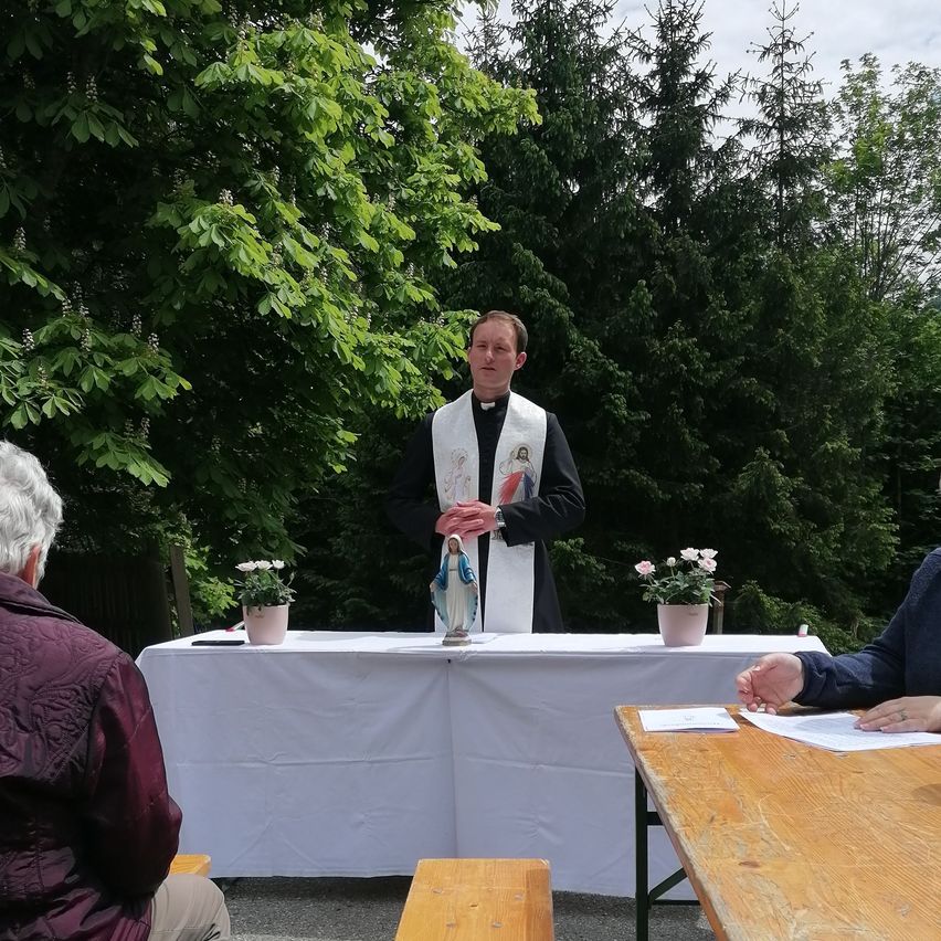 Ein Priester in einer weißen Stola hält eine Predigt im Freien. Auf dem Tisch vor ihm stehen Blumentöpfe. Menschen sitzen auf Bänken. Bäume im Hintergrund.