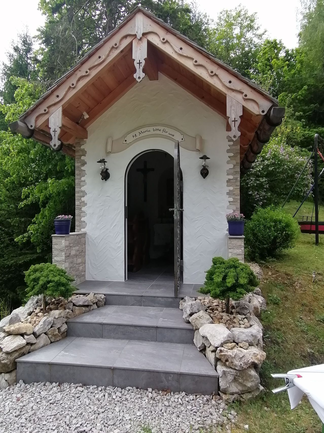 Eine kleine Kapelle mit einem Holzdach und einer Ziegelfassade. Sie hat ein Kreuz auf der Tür und zwei Lampen an den Seiten. Ein Kreuz ist auch an der Wand über der Tür. Es gibt zwei Topfpflanzen in blauen Töpfen auf beiden Seiten des Eingangs.