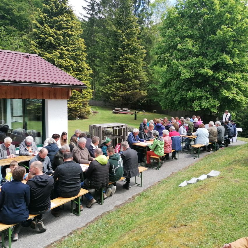 Eine Gruppe von Menschen sitzt an Tischen im Freien, möglicherweise bei einer Mahlzeit. Das Gebiet ist von Bäumen und Gras umgeben. In der Ferne ist ein Haus mit rotem Dach zu sehen.