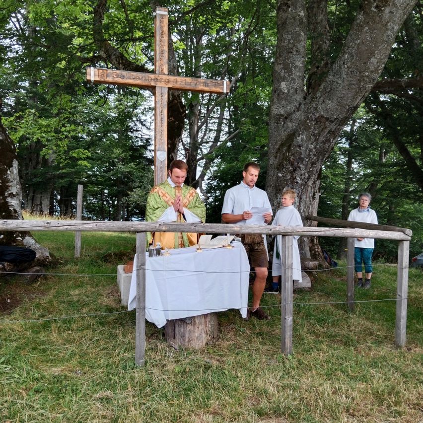 Ein Priester hält einen Gottesdienst im Freien ab, mit einem Holzkreuz und einem Altar. Zwei Männer und zwei Kinder sind anwesend und stehen hinter einem Zaun.