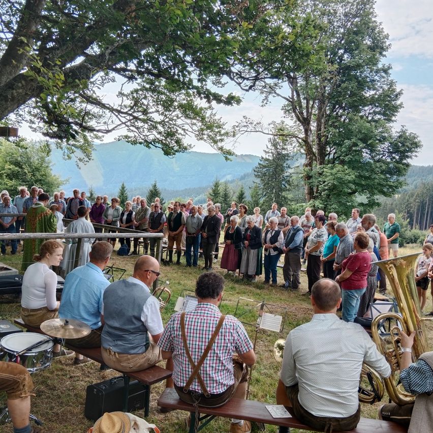 Eine große Gruppe von Menschen versammelt sich auf einer Wiese mit Bergen im Hintergrund, einige sitzen auf Bänken, andere stehen, mit Musikern, die im Vordergrund Instrumente spielen.