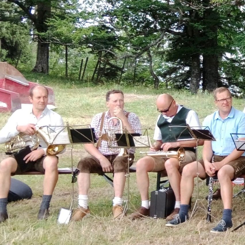 Vier Männer in traditioneller Kleidung sitzen auf einer Bank in einem Grasfeld und halten jeweils ein Musikinstrument. Sie scheinen sich auf eine Aufführung vorzubereiten.
