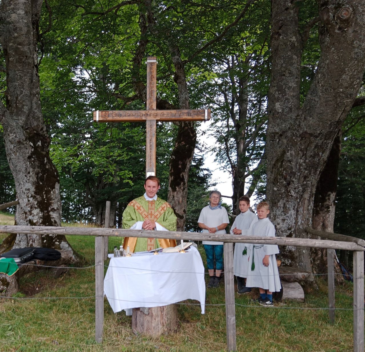 Ein Priester steht vor einem Holzkreuz im Wald, mit einem Tisch und Altartuch. Vier Kinder in weißen Gewändern stehen hinter ihm, und eine Frau steht zur Seite.