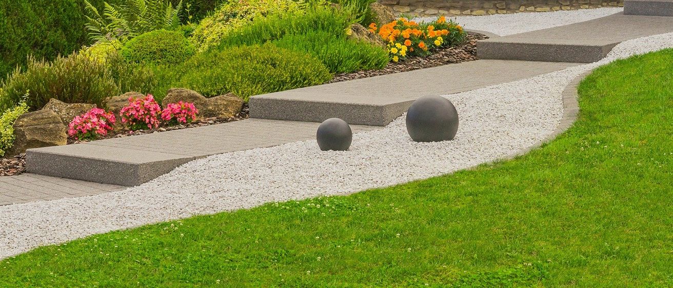 Ein Garten mit einem Kiesweg, zwei großen grauen Kugeln und verschiedenen Pflanzen und Blumen.