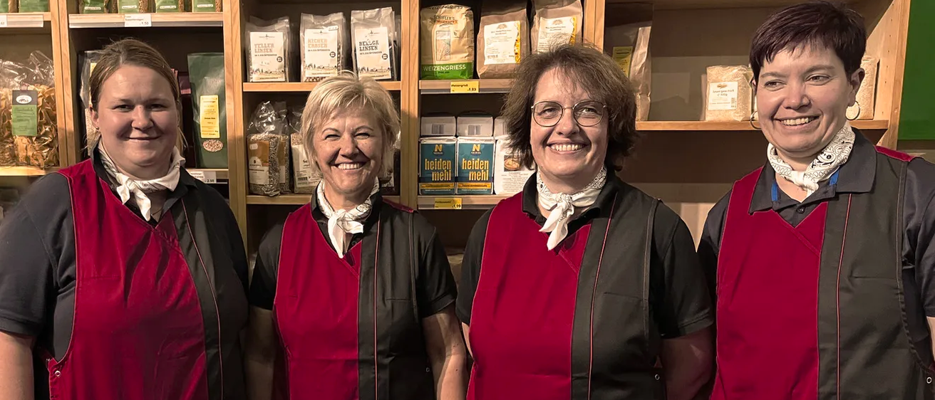 Vier Frauen in roten und schwarzen Schürzen stehen nebeneinander vor einem Holzregal, das mit verschiedenen Säcken Mehl und anderen Lebensmitteln gefüllt ist.