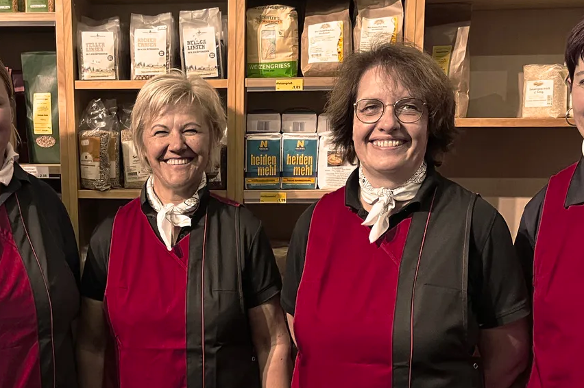 Vier Frauen in roten und schwarzen Schürzen stehen nebeneinander vor einem Holzregal, das mit verschiedenen Säcken Mehl und anderen Lebensmitteln gefüllt ist.