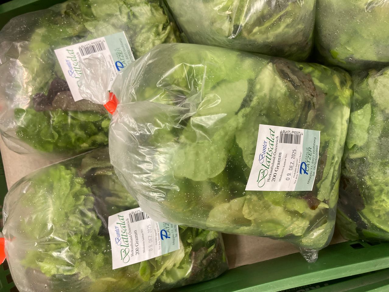 Mehrere durchsichtige Plastiktüten mit Salatblättern darin. Jeder Beutel hat ein Etikett, das darauf hinweist, dass es sich um 'Blattsalat' mit einem Gewicht von 200 Gramm handelt. Die Tüten stehen auf einem grünen Regal, und die Salatblätter sind leicht welk.