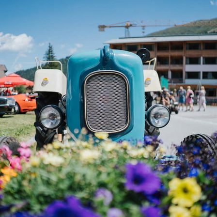 Ein blauer Vintage-Traktor ist auf einer Rasenfläche mit Blumen geparkt, mit einem Berg im Hintergrund.