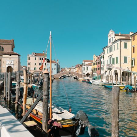Ein Kanal in Venedig mit auf beiden Seiten verankerten Booten, unter einem klaren blauen Himmel, und Gebäuden entlang des Ufers.
