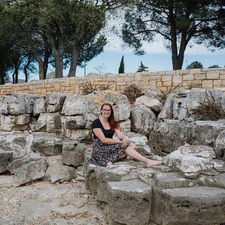 Eine Frau mit roten Haaren, die eine Brille und einen Blumenrock trägt, sitzt mit überkreuzten Beinen auf Steinstufen und blickt in die Kamera. Hinter ihr befindet sich eine Steinmauer und mehrere Bäume.