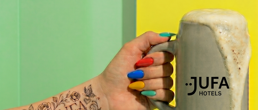 Ein tätowierter Arm mit 'Love JUFA' darauf, der einen schaumigen Bierkrug hält. Der Krug sagt JUFA HOTELS. Grüner und gelber Hintergrund.