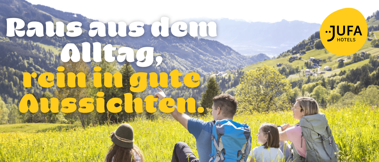 Eine Familie sitzt auf einem Grasfeld. Der Mann trägt einen Rucksack. Die Frau trägt eine Mütze. Das Mädchen sitzt neben einem Mann. Berge und Bäume sind im Hintergrund.