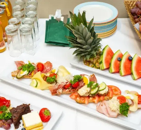 Ein Tisch zeigt verschiedene Lebensmittel auf weißen Tellern, darunter Fleisch, Käse, Obst und Gemüse. Es gibt auch Gläser mit Getränken, eine Ananas und einen Obstkorb.