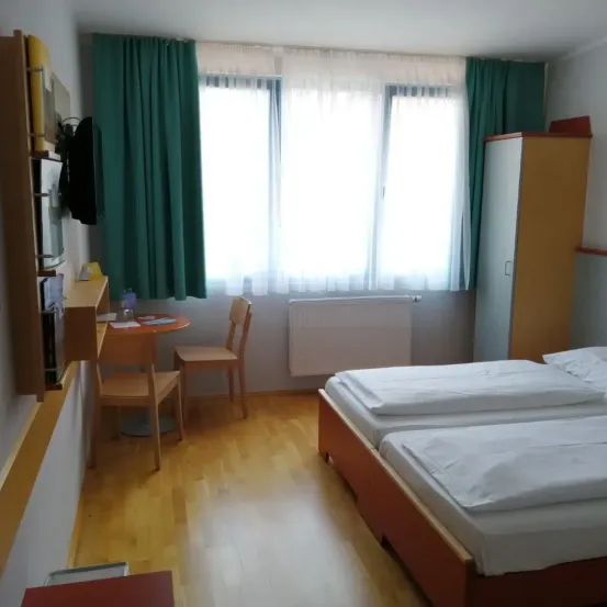 Ein Hotelzimmer mit zwei Betten, einem Schreibtisch mit Stühlen und einem Fenster mit Vorhängen. Der Raum hat Holzböden und weiße Wände.