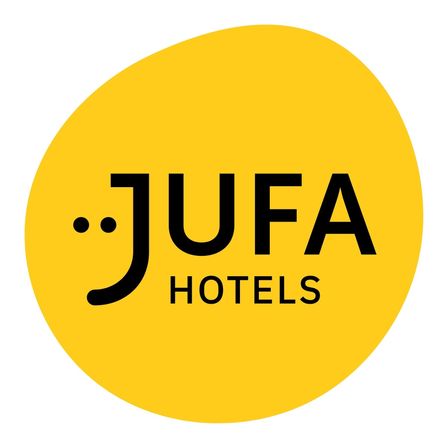 Ein gelbes kreisförmiges Logo für JUFA Hotels. Das Logo enthält den Schriftzug JUFA in stilisierter schwarzer Schrift mit einem schwarzen Punkt vor dem J. Darunter steht der Schriftzug HOTELS in normaler schwarzer Schrift.