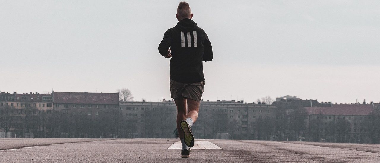Ein Mann läuft allein auf einer leeren Straße mit Gebäuden in der Ferne, trägt eine Kapuzenjacke mit drei Streifen.