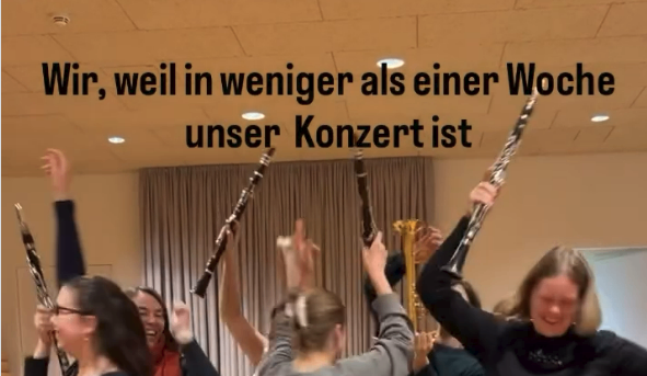 Eine Gruppe von Menschen feiert mit ihren Instrumenten und hält sie in der Luft. Der Textüberlagerung lautet: 'Wir, weil in weniger als einer Woche unser Konzert ist.' Die Umgebung ist drinnen.