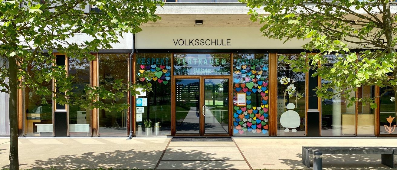 Ein modernes Gebäude mit großen Glasfenstern und einem Namensschild, das 'Volksschule' liest. Dekorative Herzen und eine Biene schmücken den Eingang, umgeben von üppigem Grün.